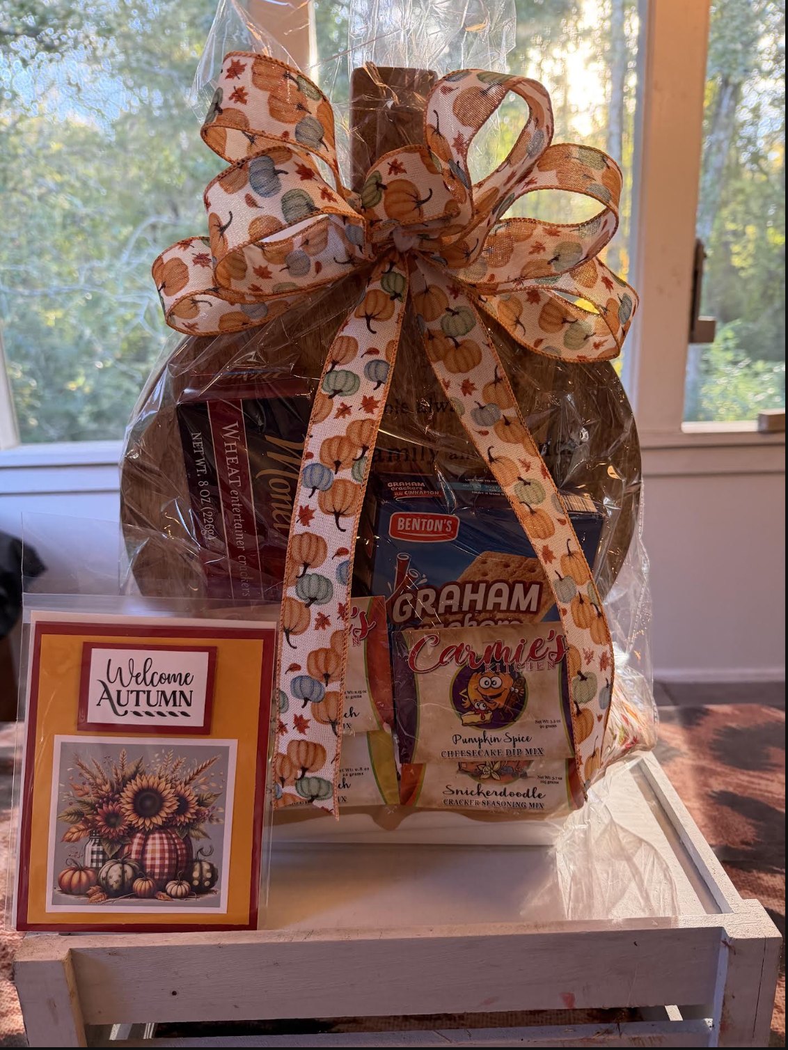 An autumn gift basket on a round wooden charcuterie board, wrapped with a pumpkin ribbon, with crackers, Carmie's Kitchen dip mixes and a Welcome Autumn card